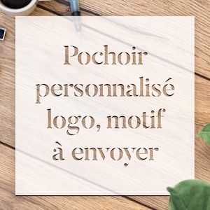 Könnte beinhalten: Eine quadratische, hellfarbene Holzschablone mit den Worten "Pochoir personnalisé logo, motif à envoyer" in einer Serifenschrift ausgeschnitten. Die Schablone liegt auf einer Holzoberfläche, im Hintergrund sind eine Tasse und ein Stift zu sehen.