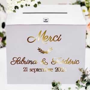 Op de afbeelding: Witte houten trouwkaartendoos met een gleuf aan de bovenkant en een kleine zilveren knop. De doos heeft gouden letters met "Merci", de namen "Sabrina & Frédéric" en de datum "21 september 2024".