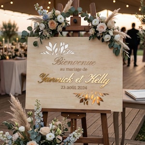 Könnte beinhalten: Holzschild zur Hochzeit mit goldfarbenem Schriftzug "Bienvenue au mariage de Yannick et Kelly 23 août 2024". Das Schild ist mit Blumenarrangements verziert und steht auf einer Holzstaffelei.