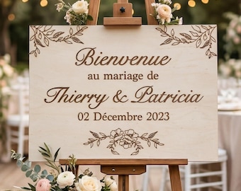 May include: A wooden wedding welcome sign on a wooden easel, decorated with flowers. The sign reads "Bienvenue au mariage de Thierry & Patricia 02 Décembre 2023". The sign has floral and leaf designs. The background is blurred with fairy lights.