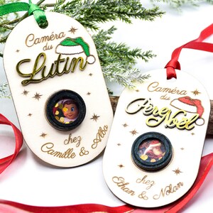 Könnte beinhalten: Zwei hölzerne Weihnachtsschmuckstücke mit einem Kameralinsen-Design. Eines trägt die Aufschrift "Caméra du Lutin" und das andere "Caméra du Père Noël". Beide haben ein kleines Foto im Inneren der Linse und sind mit Sternen verziert.