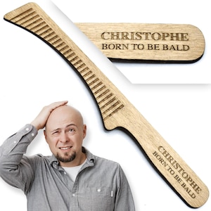 Könnte beinhalten: Ein Bartkamm aus Holz mit dem eingravierten Text "CHRISTOPHE BORN TO BE BALD". Der Kamm hat ein gebogenes Design mit gleichmäßig verteilten Zähnen. Ein kleineres Holzstück mit demselben Text ist ebenfalls sichtbar.