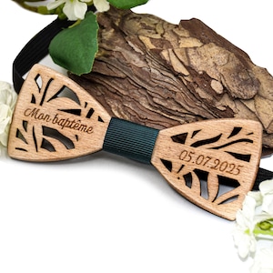 Może przedstawiać: Drewniana mucha z wycinanym laserowo wzorem i ciemnozielonym środkowym paskiem. Na muszce widnieje napis "Mon baptême" oraz data "05.07.2025". Wykonana z jasnego drewna.