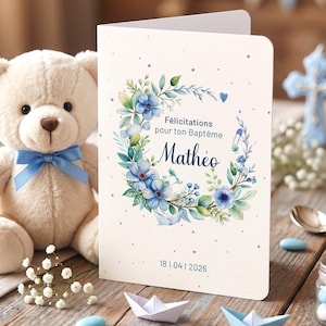 Peut inclure: Une carte de vœux pliée avec une couronne florale bleue et verte, le nom "Mathéo" et la date "18|04|2026". Un ours en peluche avec un nœud bleu et un petit bateau en papier sont au premier plan. La carte est pour un baptême.