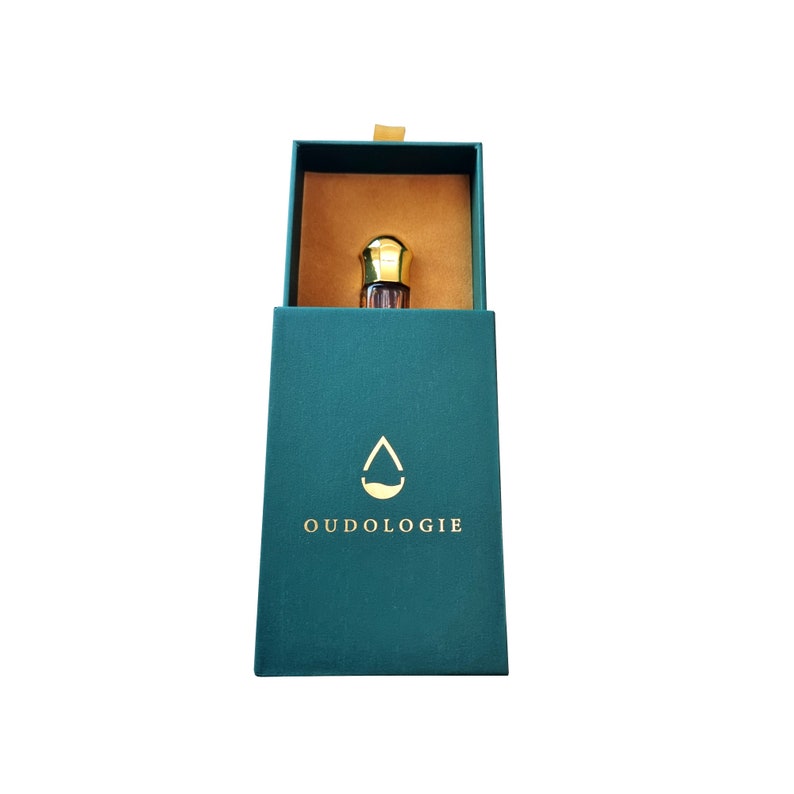 May include: A teal box with a gold interior holds a small perfume bottle with a gold cap. The box has the word "OUDOLOGIE" printed in gold, along with a gold droplet logo. The box is open, revealing the perfume bottle.