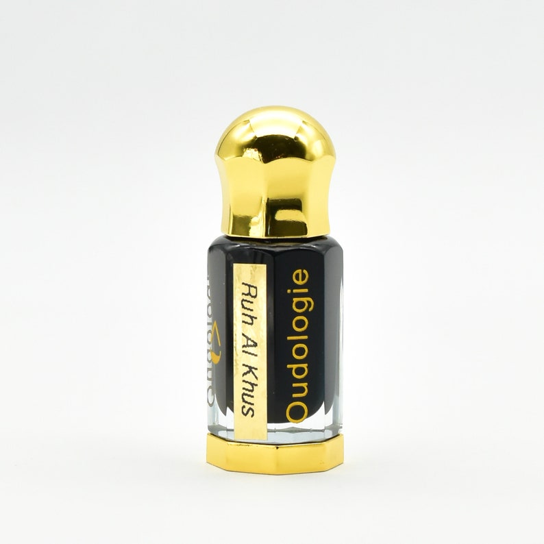 May include: A small, clear glass bottle with a gold-colored cap and base. The bottle contains a dark liquid and has a gold label with the text "Ruh Al Khus" and "Oudologie". The bottle is set against a white background.