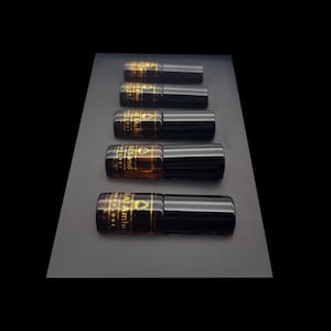 May include: Five small, dark amber glass bottles with black caps are arranged on a dark gray surface. Each bottle has a gold label with text. The bottles appear to contain a liquid, possibly perfume or essential oils.