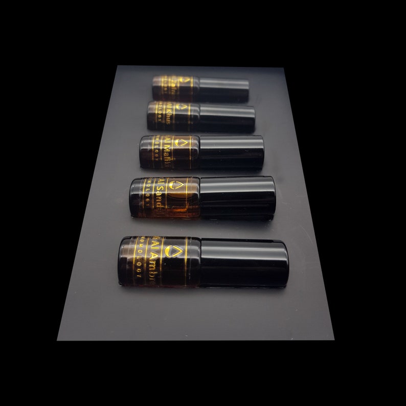 May include: Five small, dark amber glass bottles with black caps are arranged on a dark gray surface. Each bottle has a gold label with text. The bottles appear to contain a liquid, possibly perfume or essential oils.
