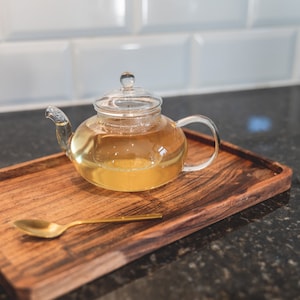 Może przedstawiać: Szklany czajnik z przezroczystą pokrywką wypełniony jasnożółtą herbatą stoi na drewnianej tacy z złotą łyżeczką.