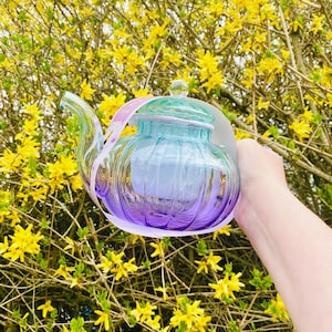 May include: A glass teapot with a purple and blue ombre design. The teapot has a clear glass lid and a handle. The teapot is being held in front of a bush of yellow flowers.