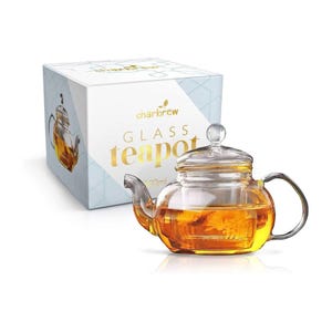 May include: A clear glass teapot with a built-in infuser, filled with yellow tea. The teapot is sitting on a white surface. The box in the background says "charbrew GLASS teapot 300ml".