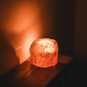 May include: A warm, orange glow emanates from a salt lamp, illuminating the surrounding area. The lamp is made of a rough, textured material and has a natural, organic shape.