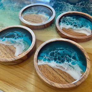 May include: Four round wooden coasters with a resin ocean wave design. The coasters feature a wooden base with a clear resin top, showcasing a turquoise and white wave pattern. The coasters are arranged on a wooden surface.