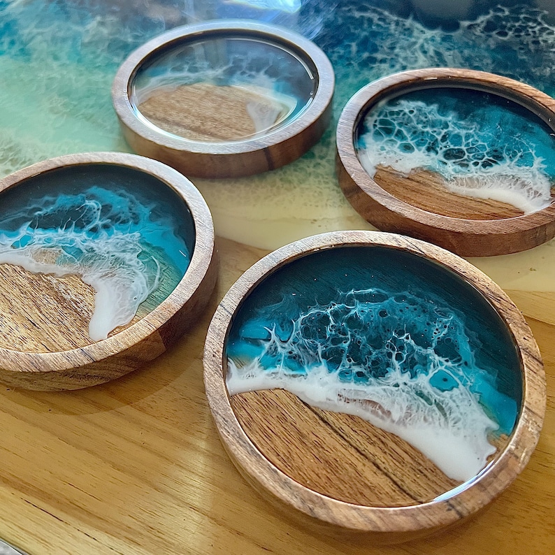 May include: Four round wooden coasters with a resin ocean wave design. The coasters feature a wooden base with a clear resin top, showcasing a turquoise and white wave pattern. The coasters are arranged on a wooden surface.
