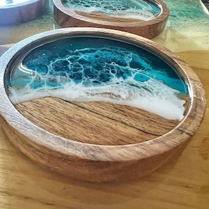 May include: Round wooden coaster with a resin design depicting a beach scene. The coaster features a wooden base with a light brown color, topped with a layer of clear resin. The resin art showcases a blue ocean with white foamy waves.