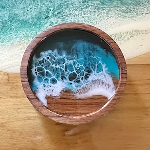 May include: A round wooden coaster with a resin ocean design. The coaster features a dark blue and turquoise ocean scene with white foamy waves. The coaster is set on a wooden surface, with a larger ocean scene in the background.