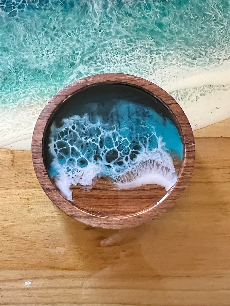 May include: A round wooden coaster with a resin ocean design. The coaster features a dark blue and turquoise ocean scene with white foamy waves. The coaster is set on a wooden surface, with a larger ocean scene in the background.