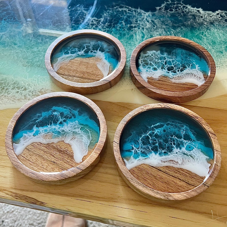 May include: Four round wooden coasters with a beach scene design. The coasters feature a wooden base with a resin top depicting a blue ocean and white waves crashing on a sandy shore. The coasters are arranged on a wooden surface.