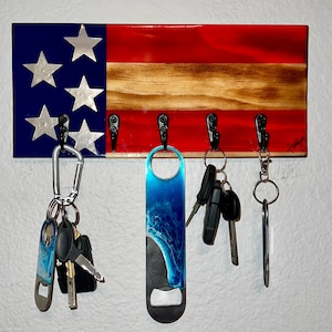 May include: A wooden key holder shaped like an American flag with five silver stars on a blue background, red and white stripes, and five black hooks. A bottle opener with a blue and white wave design, a carabiner, and a set of keys are hanging from the hooks.