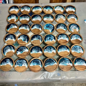 May include: A collection of wooden coasters with a resin ocean wave design. Each coaster features a wooden base with a sandy beach and a blue and white wave pattern. The coasters are arranged on a glass surface.