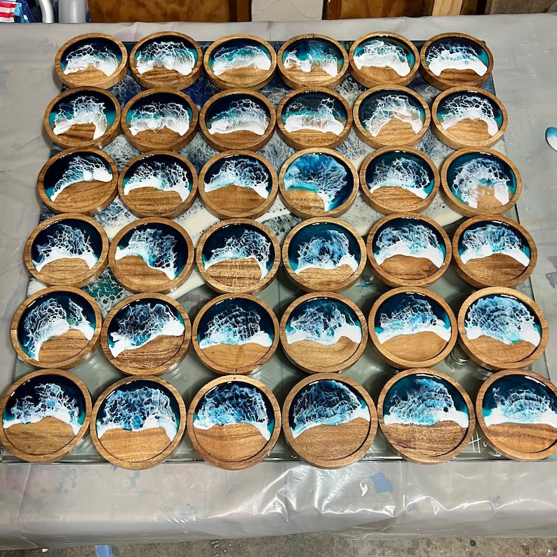 May include: A collection of wooden coasters with a resin ocean wave design. Each coaster features a wooden base with a sandy beach and a blue and white wave pattern. The coasters are arranged on a glass surface.