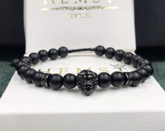 May include: A beaded bracelet featuring matte black spherical beads, two dark metallic spacers with small embedded stones, and a central black lion head charm. The bracelet is displayed in front of a white box with the text HEMST JEWELRY.