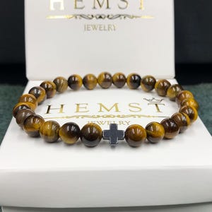 May include: A brown tiger's eye beaded bracelet with a black metal cross charm. The bracelet is on a white box with the text "Hemst Jewellery" printed on it.