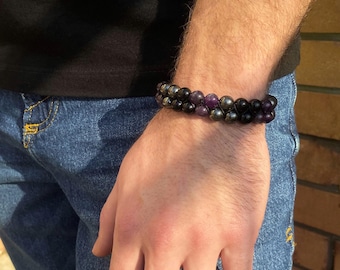 May include: A double-strand bracelet featuring black and purple beads. The bracelet is crafted from natural stones and is worn on a person's wrist.