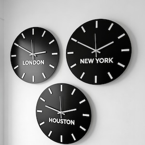 May include: Three circular black wall clocks with white hour markers and white hands. Each clock features a white city name printed below the center: London, New York, and Houston. The clocks are arranged in a cluster on a plain white wall surface.