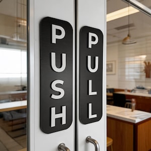 May include: Black and white door signs with the words "PUSH" and "PULL" in bold, white letters. The signs are rectangular with rounded corners and are mounted on a white door. Silver door handles are visible below the signs.