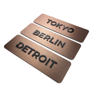 May include: Three rectangular metal signs with a brushed copper finish. The signs are stacked on top of each other and feature the words "DETROIT", "BERLIN", and "TOKYO" in black lettering.