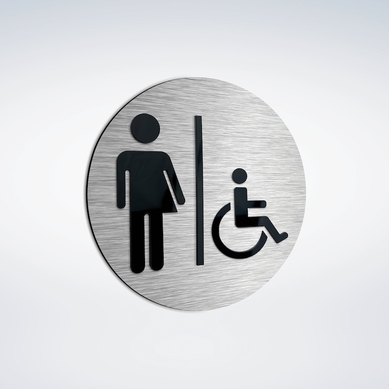 May include: A round, brushed silver sign with black symbols. The sign shows a stick figure of a person with a skirt, a vertical line, and a person in a wheelchair.