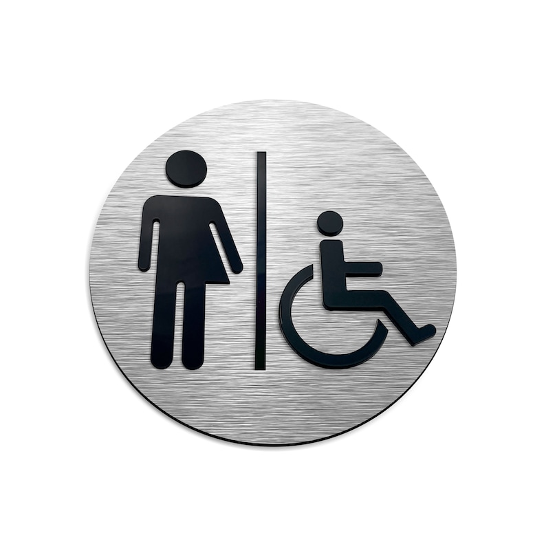 May include: A round, brushed silver sign with black symbols. The sign features a gender-neutral stick figure with a skirt, a vertical line, and a person in a wheelchair.