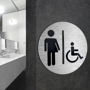 May include: A silver round sign with black symbols. The sign shows a person and a wheelchair symbol. The sign is mounted on a gray wall.
