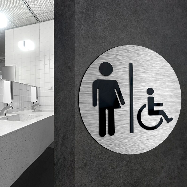 May include: A silver round sign with black symbols. The sign shows a person and a wheelchair symbol. The sign is mounted on a gray wall.