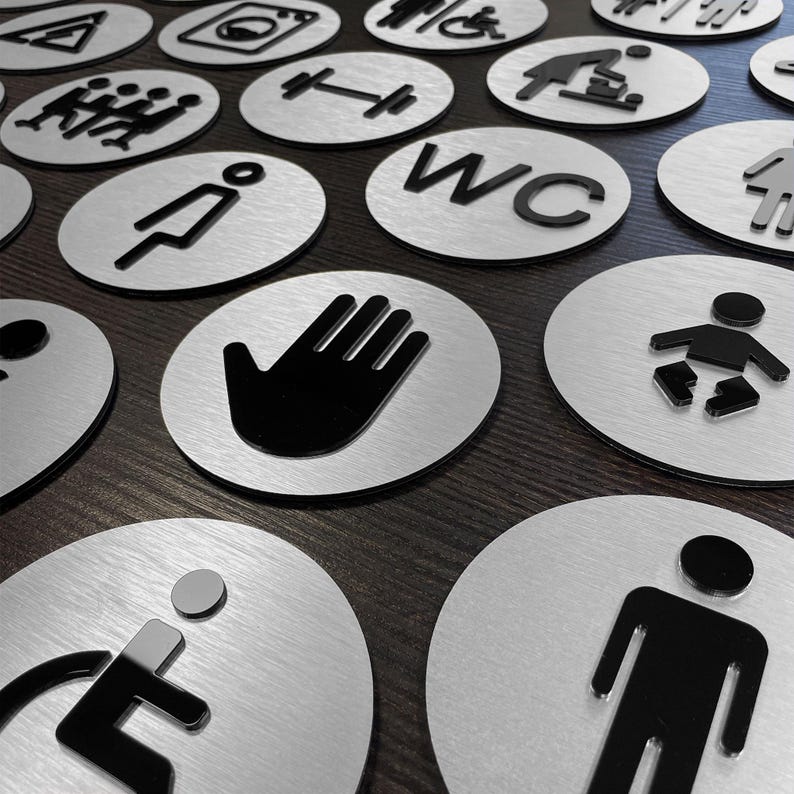 May include: A collection of round, silver metal signs with black symbols. The signs feature various symbols, including a wheelchair, a toilet, a hand, a baby, and a person.