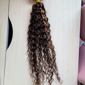 May include: A set of brown and blonde curly hair extensions with micro rings attached. The hair is arranged in a long strand.