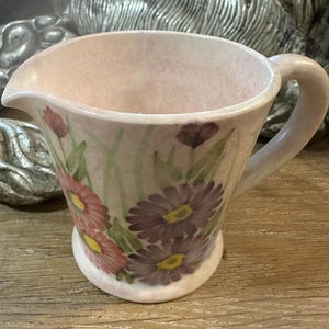 May include: A pink ceramic jug with a floral design featuring purple and pink flowers. The jug has a handle and a rounded base.