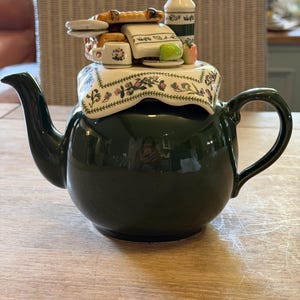Puede incluir: Una tetera de cerámica verde oscuro con una tapa que presenta una escena de picnic en miniatura. La tapa está cubierta por un paño blanco con borde floral, rematado con una cesta pequeña, una botella blanca, una taza diminuta y una manzana.