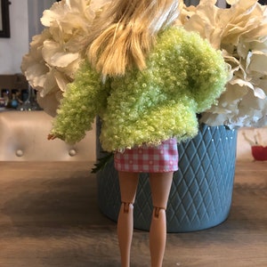 Puede incluir: Una muñeca con una chaqueta verde peluda y una falda a cuadros rosa y blanca. La muñeca tiene el pelo rubio y está de pie frente a un jarrón de flores blancas.