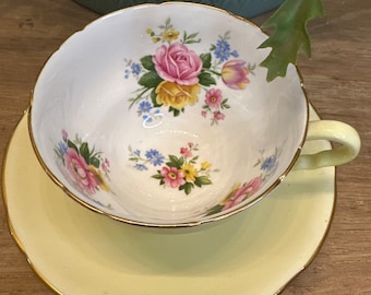 May include: A vintage teacup and saucer set with a floral pattern. The teacup has a white background with pink, yellow, and blue flowers. The saucer is a pale yellow color with a gold rim.