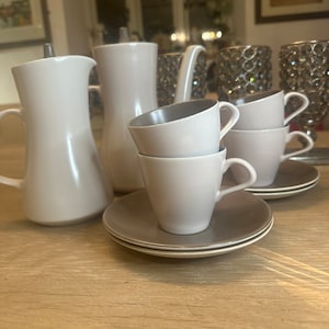 Puede incluir: Un juego de tazas de té y platillos de cerámica marrón claro con una tetera y una jarra de crema a juego. Las tazas y los platillos tienen un diseño simple y elegante.