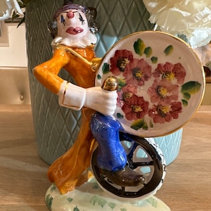 Könnte beinhalten: Eine Keramikfigur eines Clowns, der ein Einrad fährt. Der Clown trägt ein buntes Kostüm und einen Hut. Das Einrad hat ein Rad mit Blumenmuster.