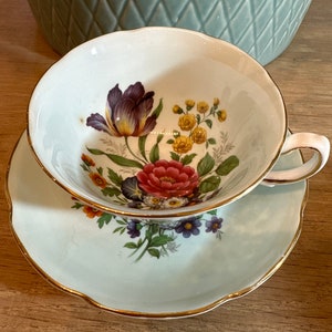 Op de afbeelding: Een vintage theekopje en schotelset met een bloemenmotief. Het kopje en de schotel zijn beide wit met een lichtblauwe rand en een gouden rand. Het bloemenmotief bevat een verscheidenheid aan bloemen in tinten roze, rood, geel en paars.