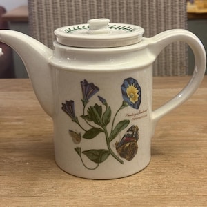 Könnte beinhalten: Cremefarbene Keramik-Teekanne mit Deckel und grünem botanischen Design. Die Teekanne ist mit Illustrationen von blauen und lila Blumen, grünen Blättern und einem Schmetterling verziert. Der Text "Trailing Bindweed Convolvulus" ist ebenfalls sichtbar.