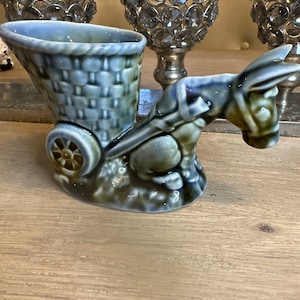 May include: A blue and green ceramic donkey pulling a small cart with a basket. The donkey has a saddle and a bridle. The cart has a wheel and a basket. The donkey and cart are on a wooden surface.