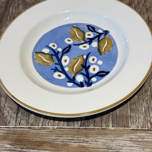 May include: A white ceramic plate with a blue rim and a blue centre featuring a stylised floral design with white and gold accents.
