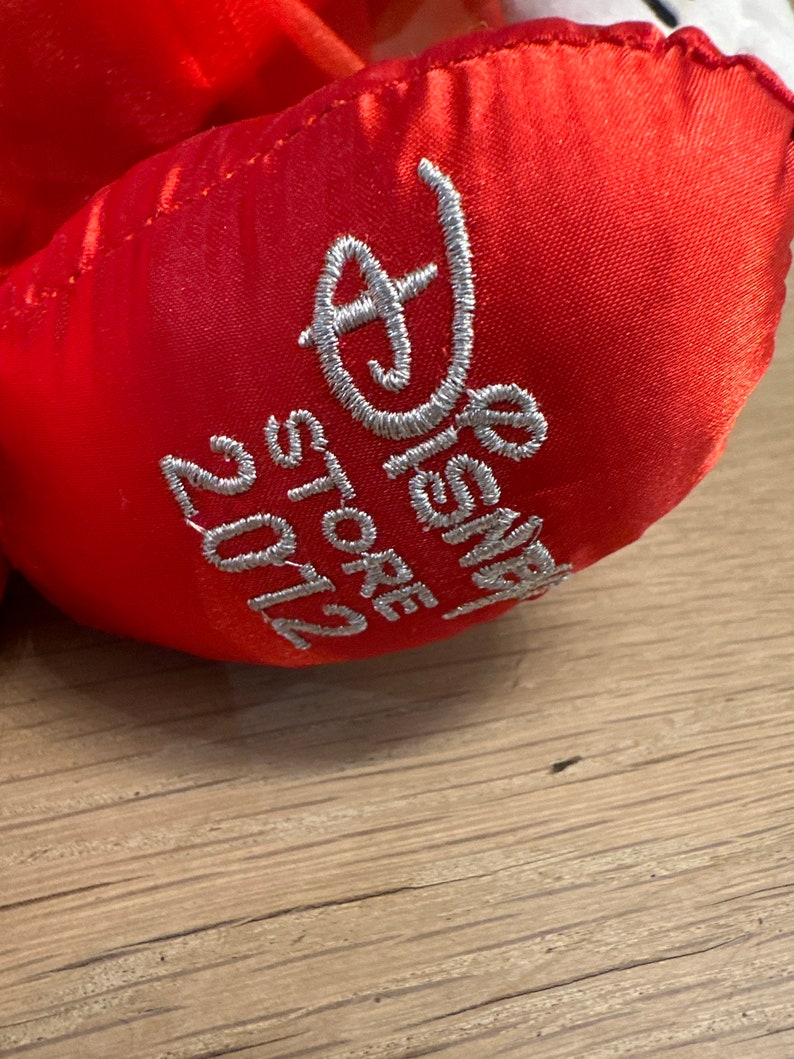 K&ouml;nnte beinhalten: Nahaufnahme eines roten Pl&uuml;schtierfu&szlig;es mit silbernem, gesticktem Text. Der Text lautet "Disney Store 2012". Das Pl&uuml;schtier befindet sich auf einer Holzoberfl&auml;che.