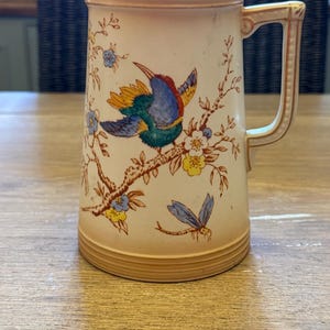 Puede incluir: Jarra de color crema con un diseño de pájaro y flores. El pájaro tiene plumas azules, amarillas y verdes, posado en una rama marrón con flores azules y amarillas. La jarra tiene un borde y un asa dorados.