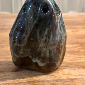 Puede incluir: Un pequeño jarrón de cerámica de color verde oscuro y marrón con una forma única e irregular. El jarrón tiene varios agujeros alrededor de la parte superior, lo que lo hace adecuado para sostener pequeñas flores o arreglos secos.